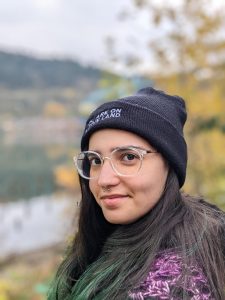 Image of a person wearing a black beanie and a purple shawl, the backgroudn is blurred, the person is wearing glasses and is smiling. 